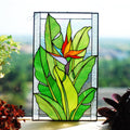 Handmade stained glass Bird of Paradise panel with orange flower and green leaves, hanging in a sunny window, Tiffany copper foil technique