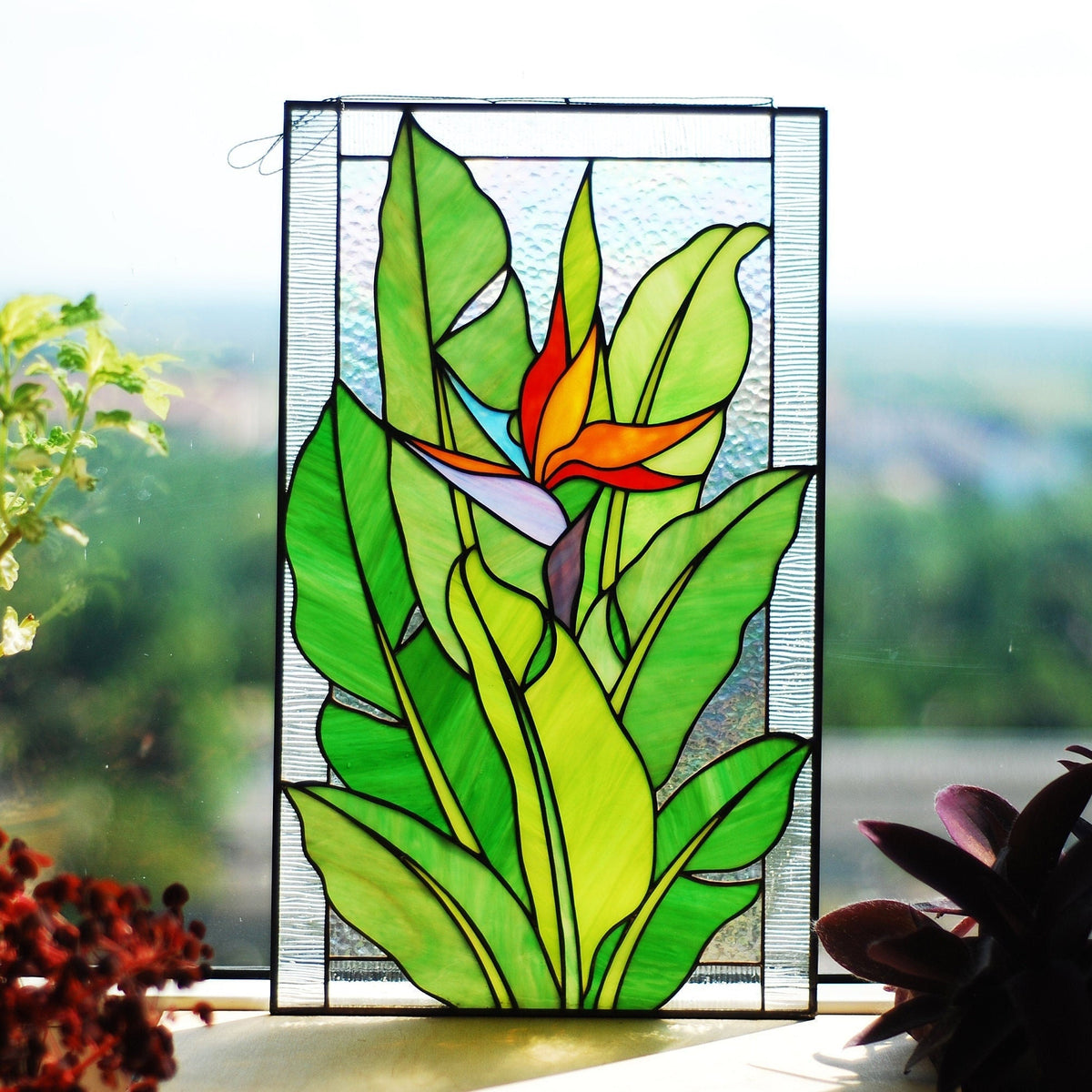 Handmade stained glass Bird of Paradise panel with orange flower and green leaves, hanging in a sunny window, Tiffany copper foil technique