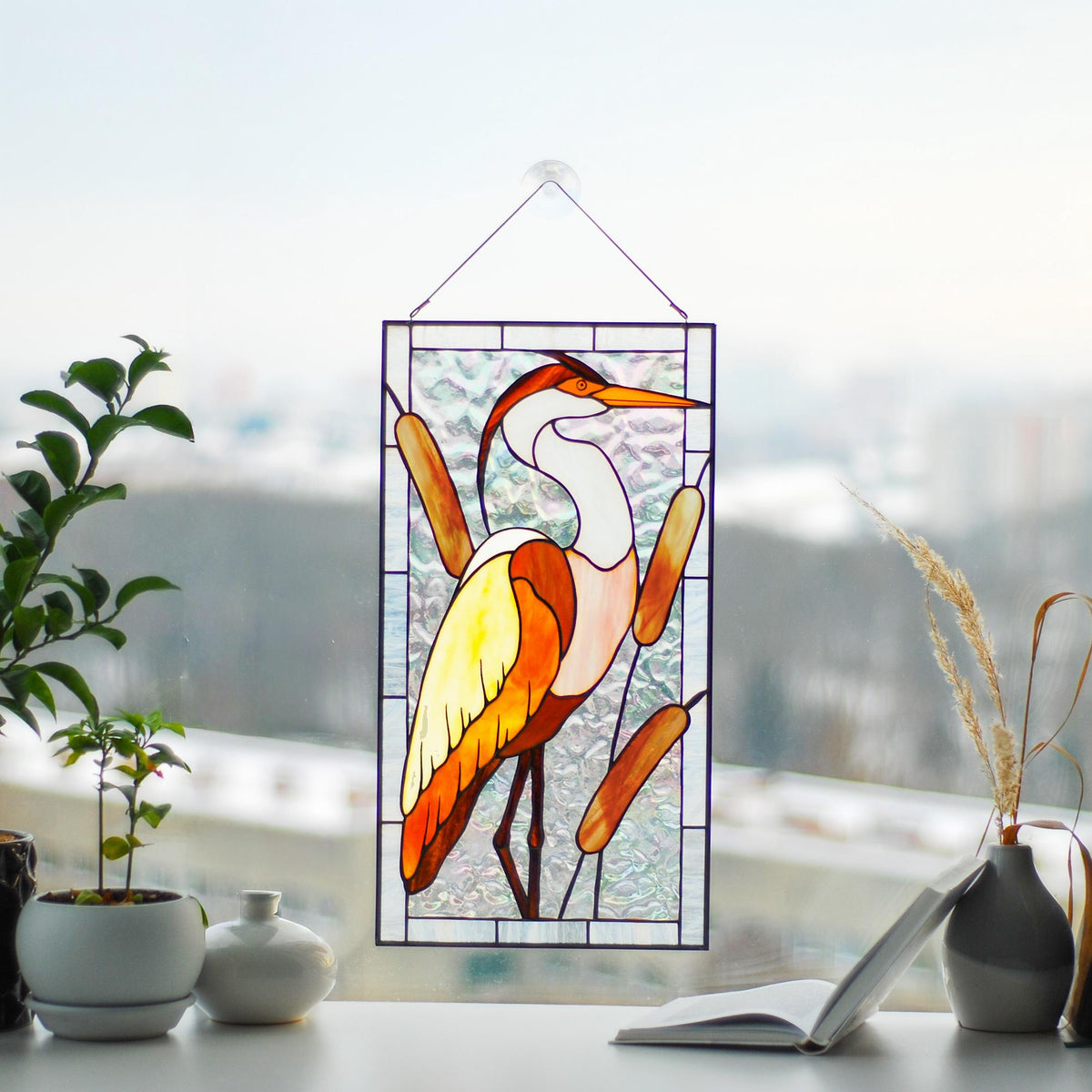 Stained glass heron and reeds window panel displayed on a windowsill with indoor plants