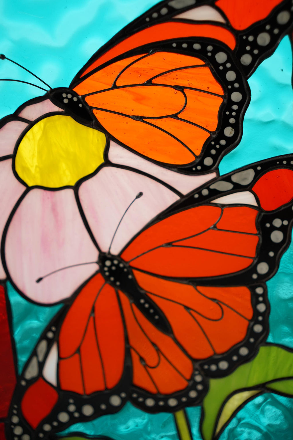 Close-up of stained glass monarch butterfly on pink flower, vibrant orange and turquoise cathedral glass