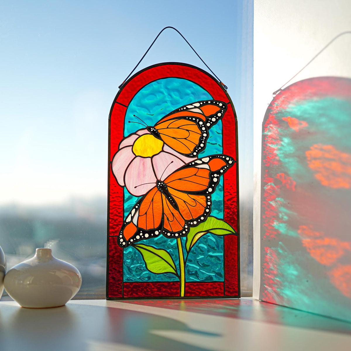 Stained glass monarch butterfly panel hanging in a bright window, red arch frame, turquoise background