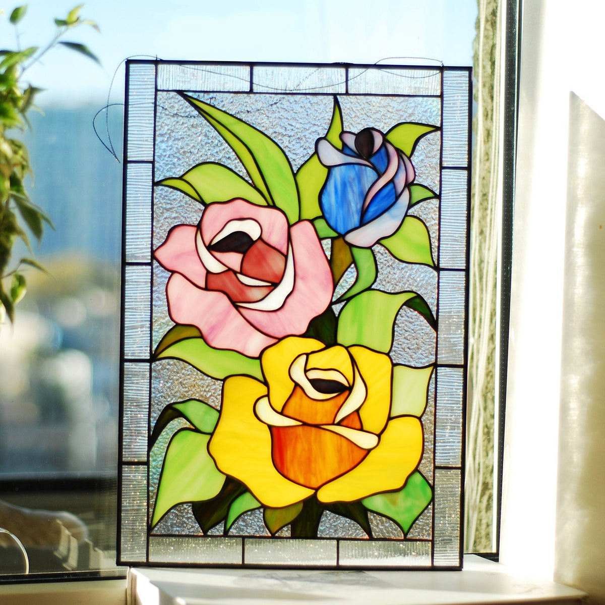 Stained glass roses panel with pink, yellow and blue roses, displayed on a bright windowsill with plants