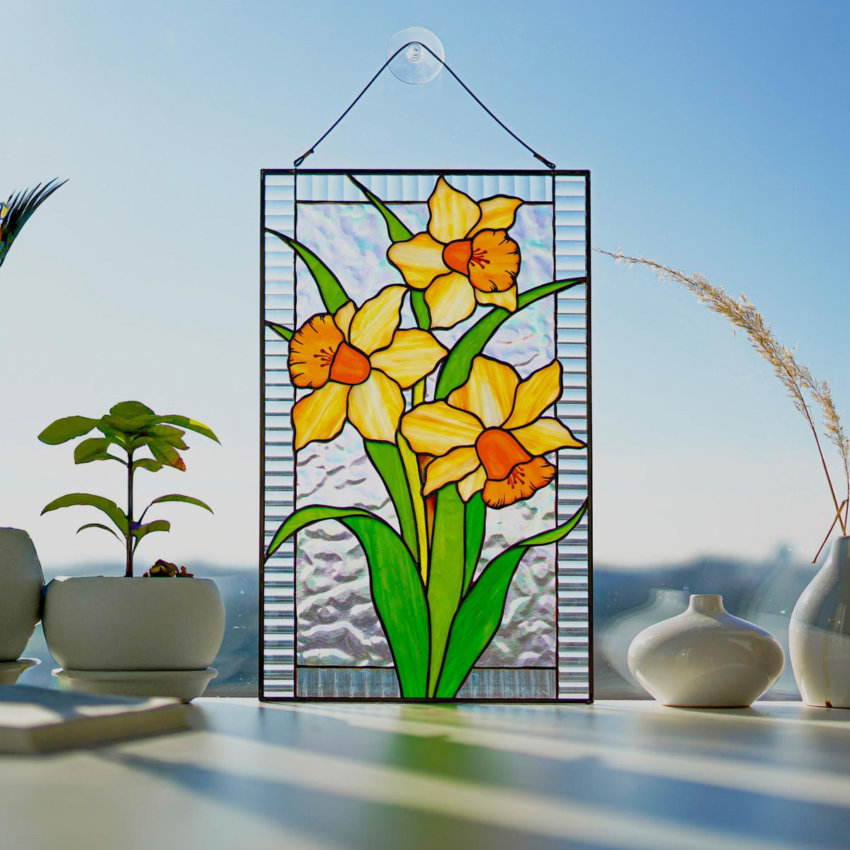 Stained glass yellow orange narcissus panel with iridescent background, displayed on a bright windowsill with vases