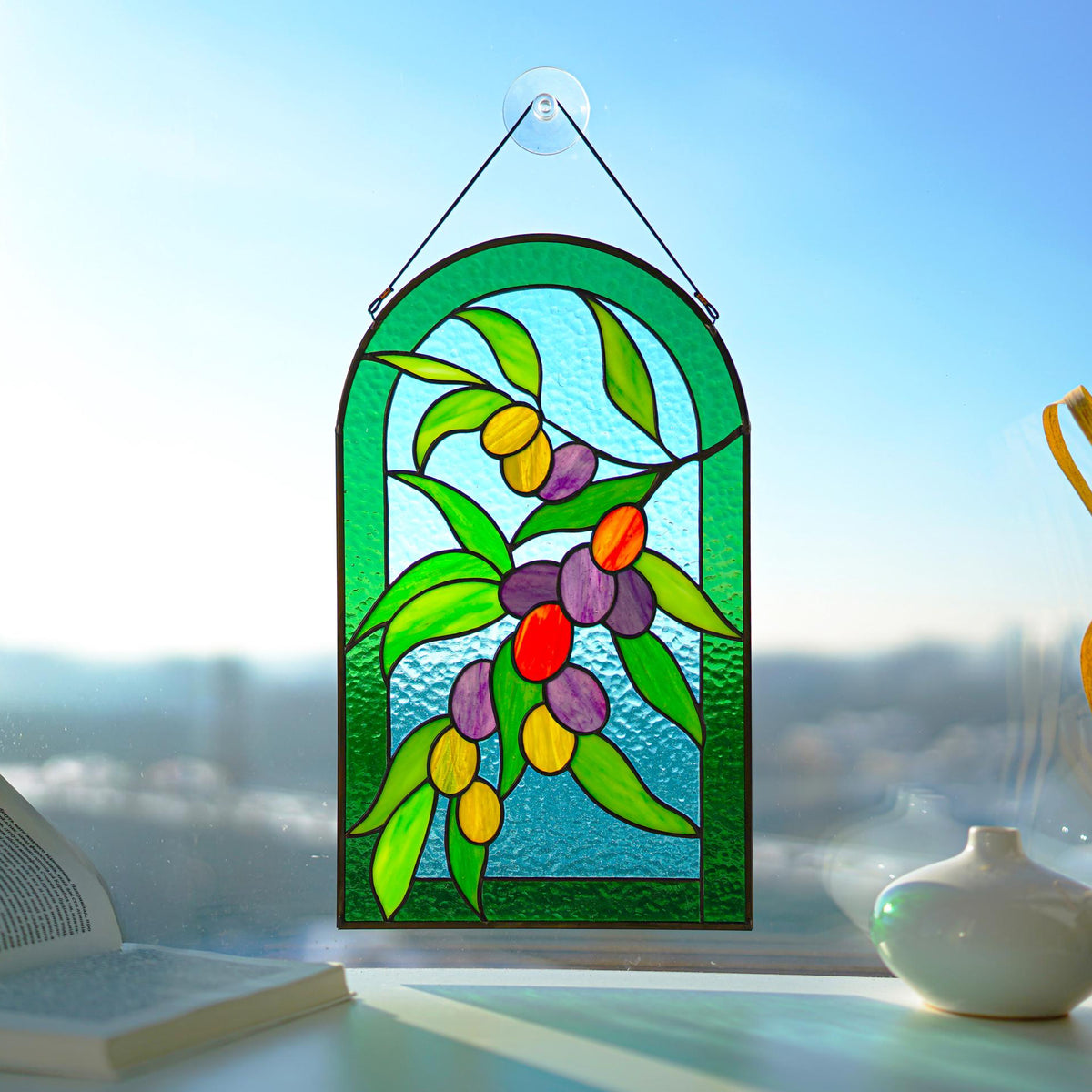 Stained glass olive branch panel with green leaves and colorful olives, displayed on a bright windowsill