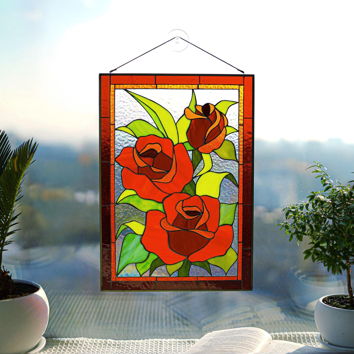 Stained glass red rose panel with red zinc frame, displayed on a bright windowsill with indoor plants