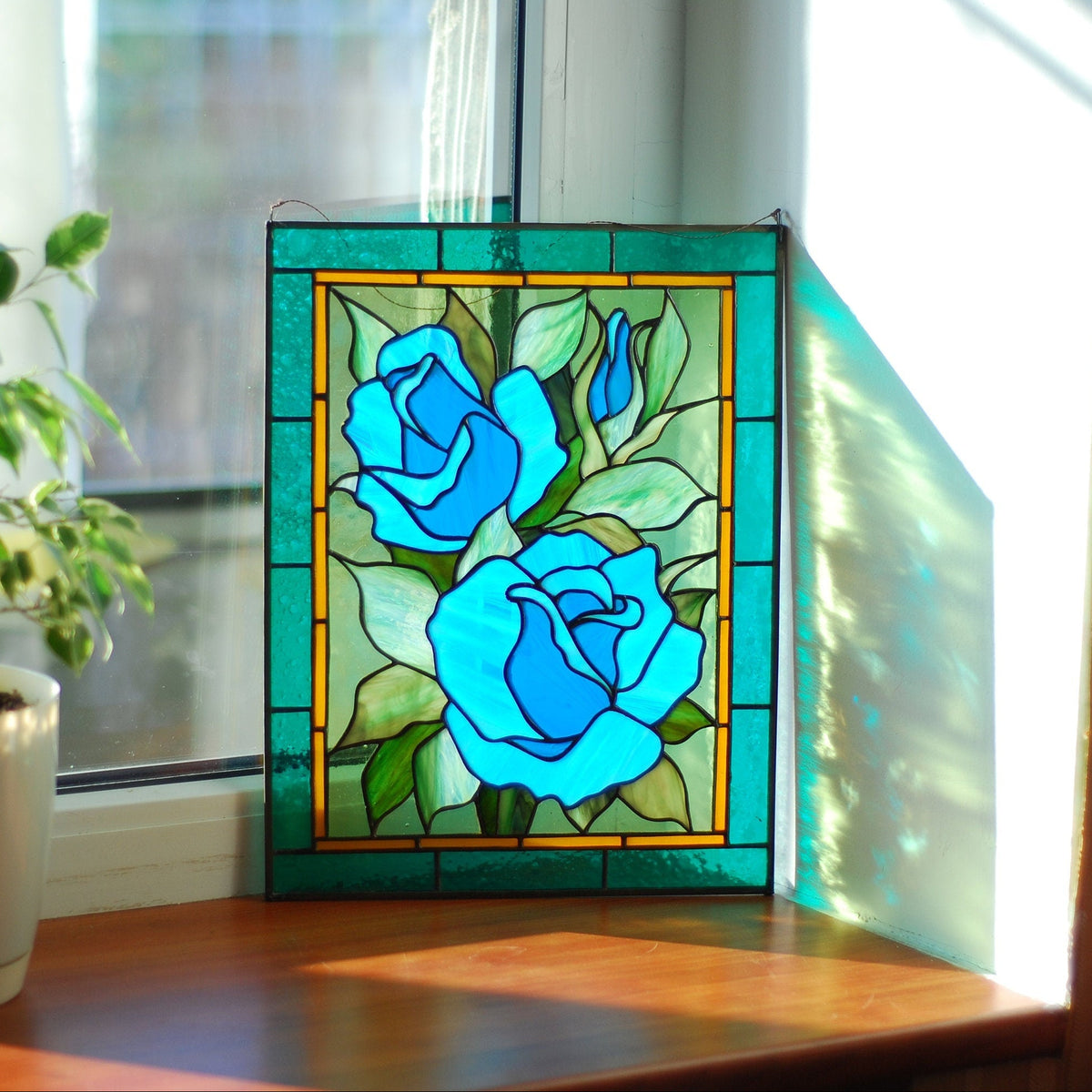 Stained glass blue rose window panel displayed in a home interior with plants, jewel-toned turquoise floral art, handmade Tiffany style
