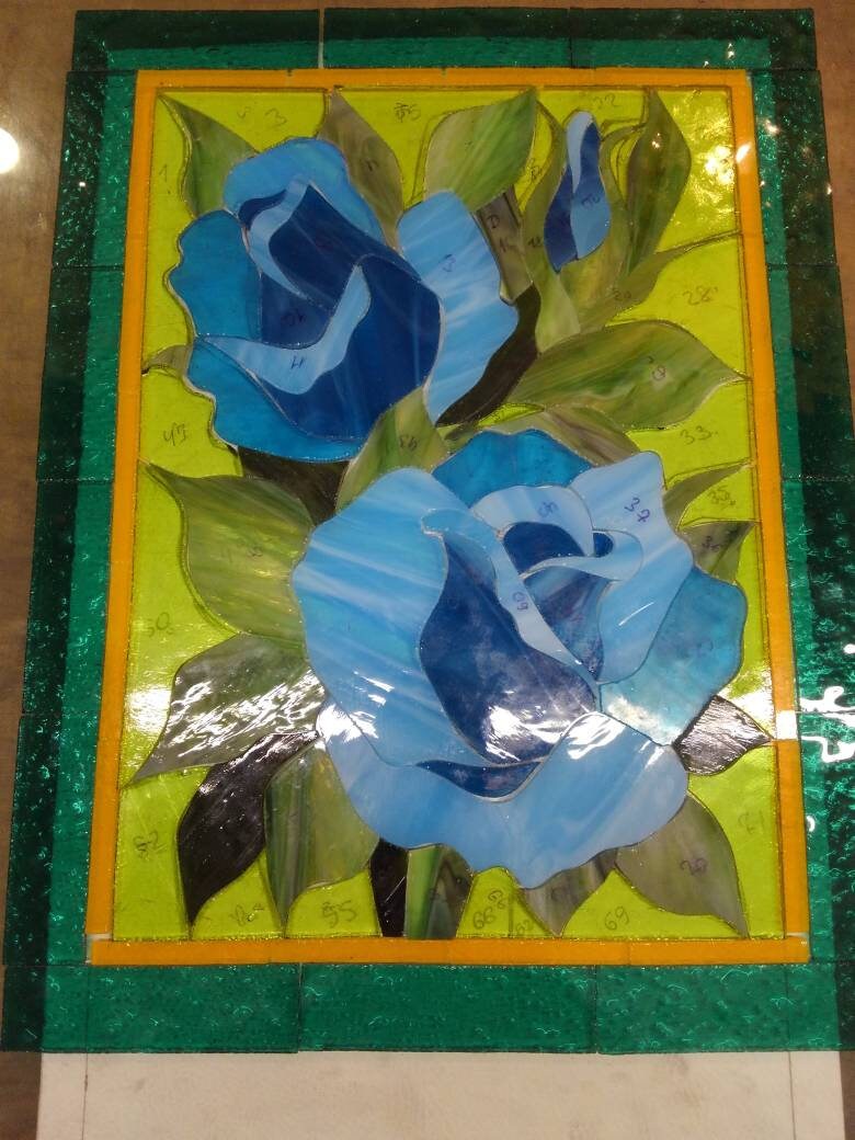 Handcrafted stained glass blue rose panel during production, copper foil and soldering process, turquoise floral window art in progress
