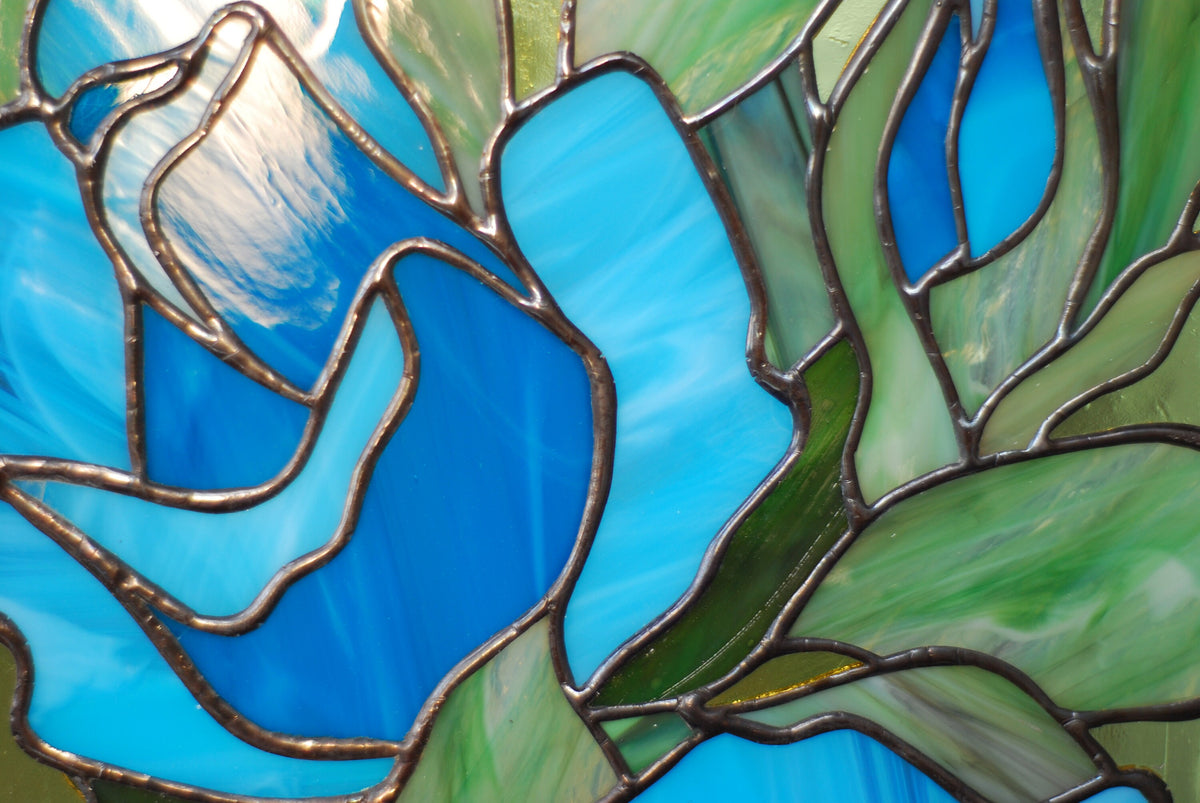 Close-up of stained glass blue rose panel showing copper foil soldering detail, teal and turquoise cathedral glass with green background
