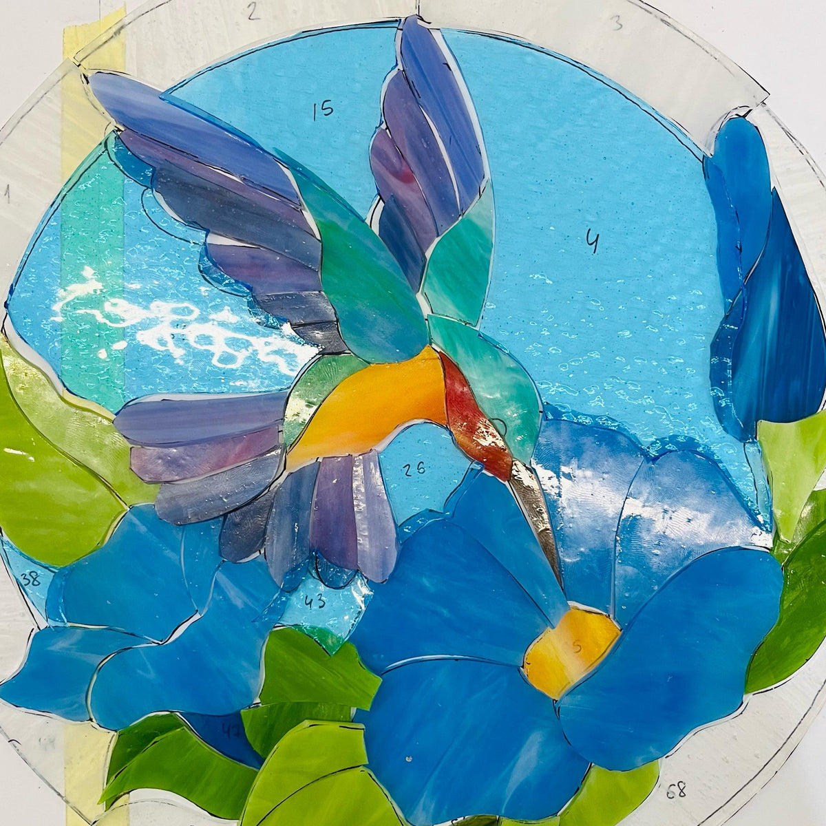 Stained glass hummingbird panel work in progress on a light table, blue and green glass pieces with copper foil technique, handmade process
