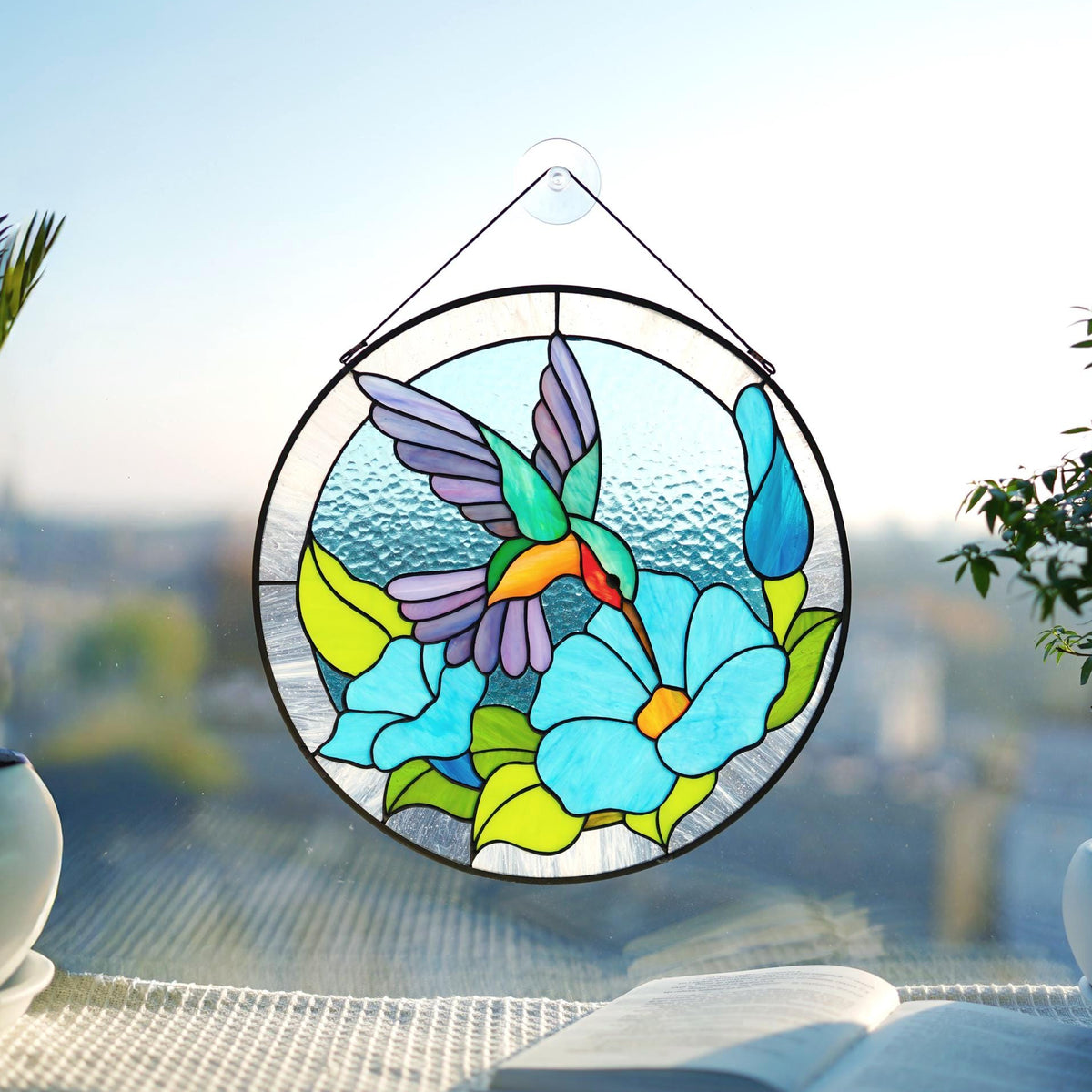 Handmade round stained glass hummingbird suncatcher on a sunny terrace with plants, teal and blue morning glory flowers, Tiffany copper foil, 12 inches
