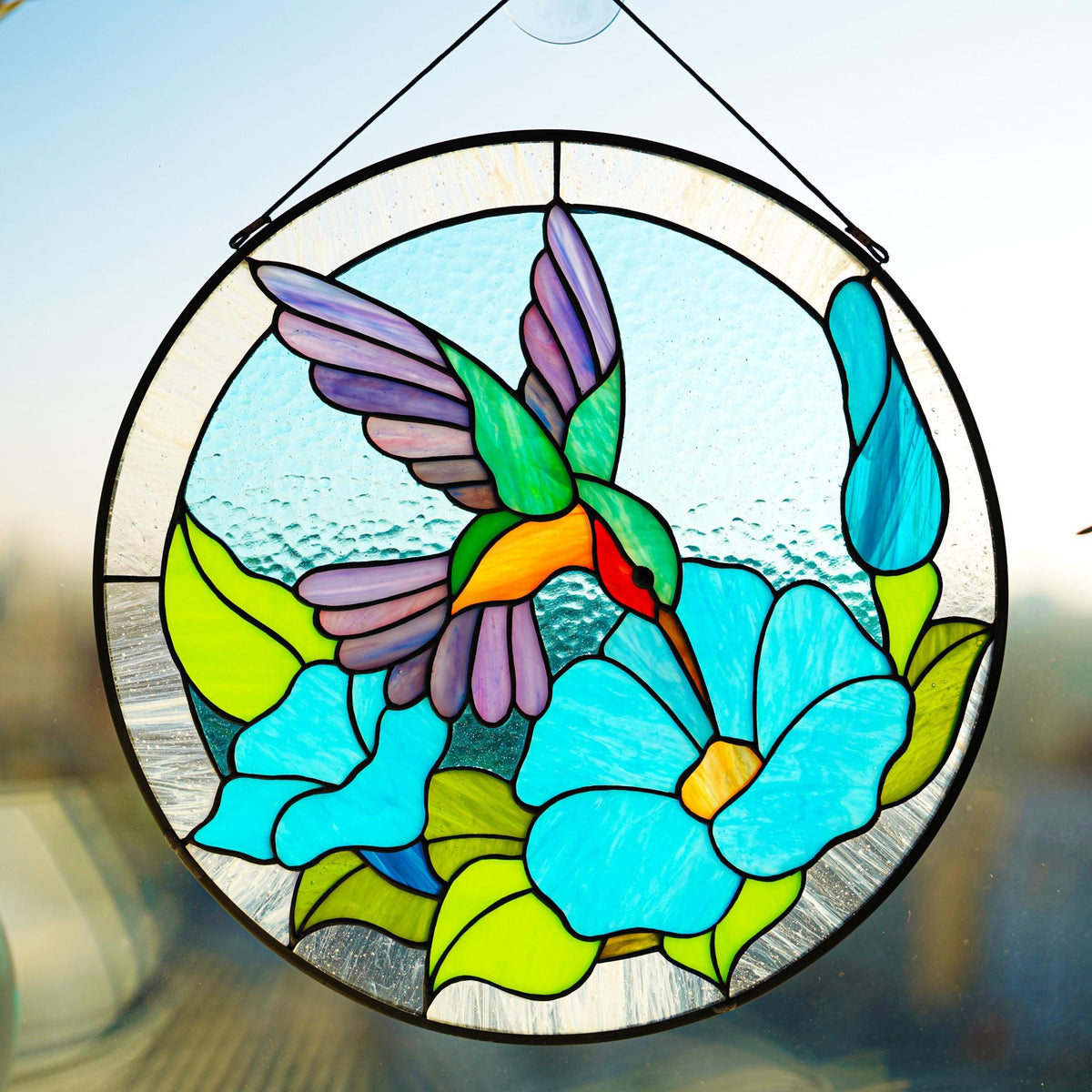 Round stained glass hummingbird suncatcher hanging in a window with natural light shining through, teal and blue cathedral glass glowing
