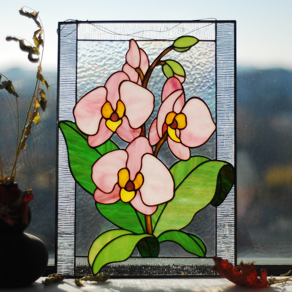 Stained glass orchid panel displayed on a windowsill with outdoor light shining through, translucent pink and white cathedral glass glowing

